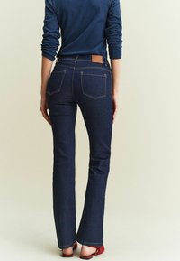 Jean évasé bleu foncé avec une texture lisse, comportant des poches arrière et un patch en cuir brun sur la taille. Porté avec des chaussures plates rouges.