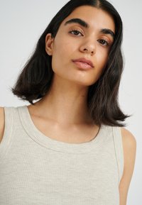 Light beige ribbed tank top with a round neckline, featuring a textured fabric and a fitted design. Hair is dark and slightly wavy.