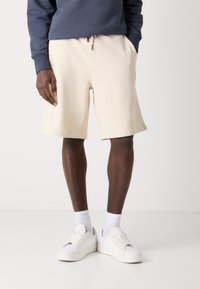 Beige cotton shorts with a relaxed fit, featuring an elastic waistband, drawstring, and side pockets, paired with white sneakers and socks.