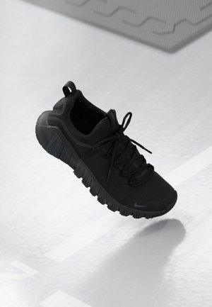 Czarny sportowy sneaker z grubą podeszwą i sznurowadłami, zawieszony nad jasną, teksturowaną podłogą z cieniem poniżej.