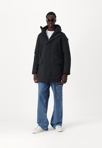 Parka nera con tasche frontali, chiusura con zip e cappuccio regolabile. Indossato con jeans blu larghi e sneaker bianche.