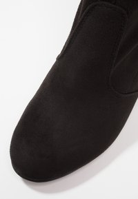 Gros plan sur l'avant d'une chaussure en daim noir avec des coutures visibles et une forme lisse et arrondie sur fond blanc.