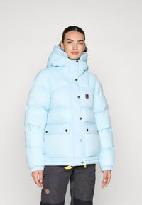 Lichtblauwe pufferjack met een hoge kraag, voorzien van een logopatch, drukknoopsluitingen en twee grote voorzakken. Zachte, gequiltte textuur.