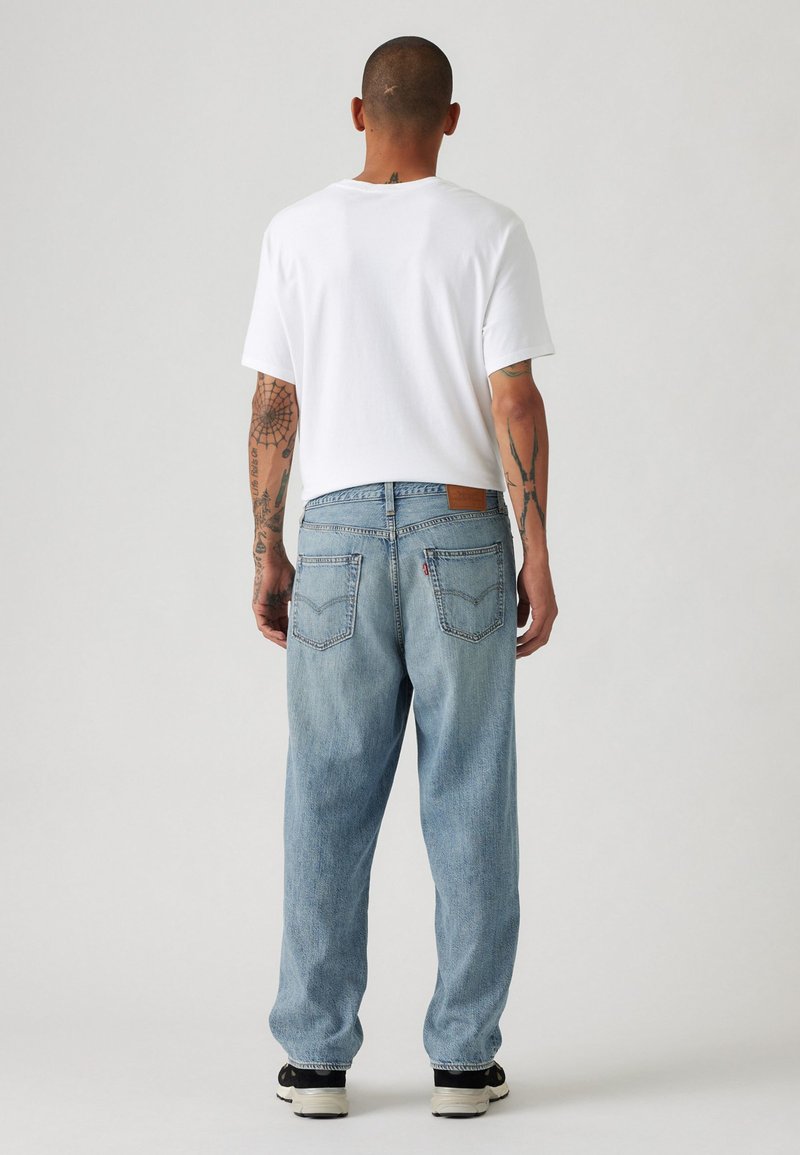Lichtblauwe denim jeans met een relaxte pasvorm, vijf zakken ontwerp en een bruine leren patch op de achterkant van de tailleband. Witte t-shirt gedragen.