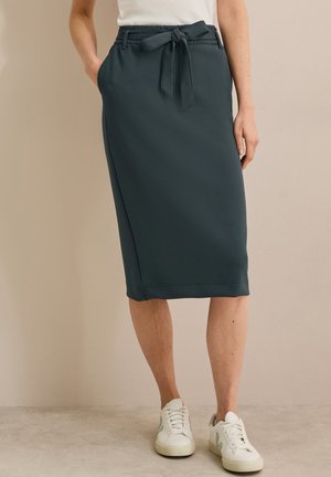 Jupe midi teal foncé avec taille élastique, détail de ceinture nouée, poches latérales et texture de tissu lisse. Associée à des baskets blanches.
