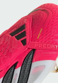 Κοντινή λήψη του κόκκινου ποδοσφαιρικού παπουτσιού Adidas Predator που δείχνει το μαύρο λογότυπο, το κείμενο "NANOSTRIKE+", υφή από υφασμάτινο υλικό και μαύρο σχεδιασμό με μοτίβο στη σόλα.