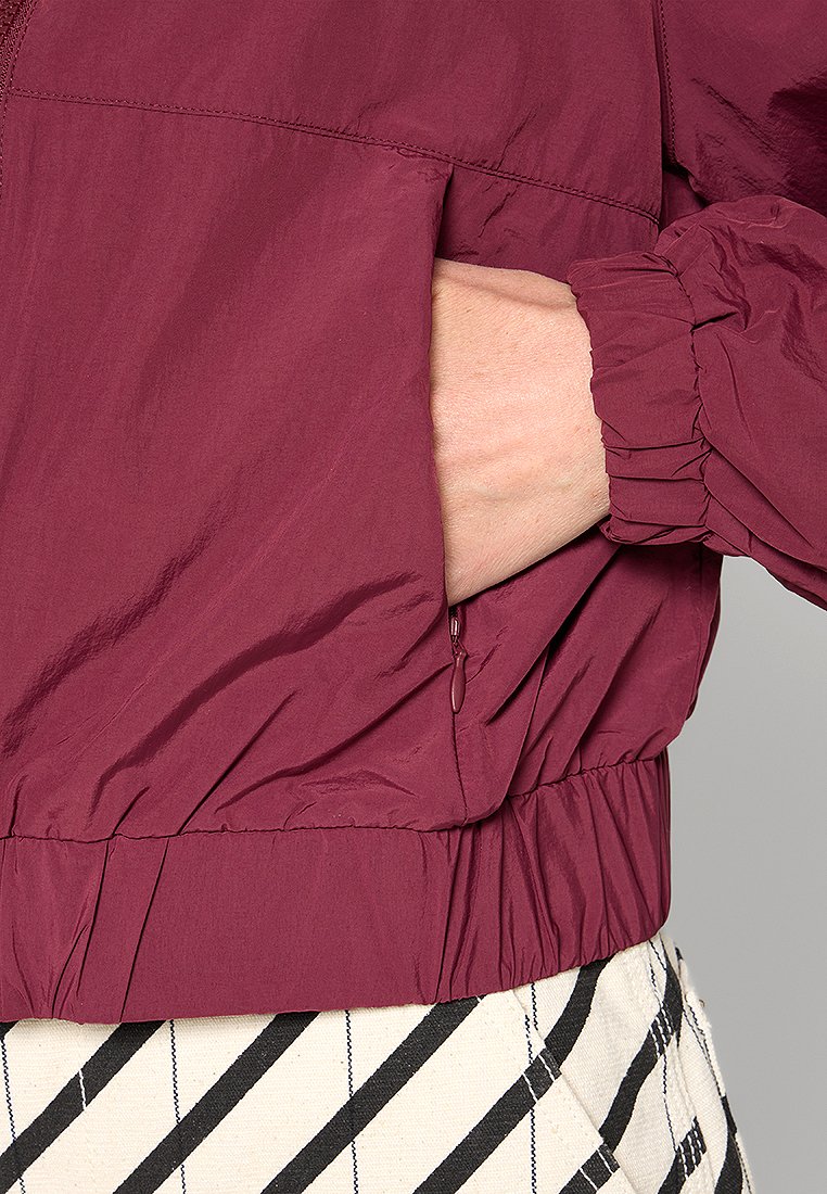 Bordeaux kleurige lichtgewicht jas met elastische manchetten en tailleband. Voorzien van een zijzak met rits. Draagt over een gestreepte rok.