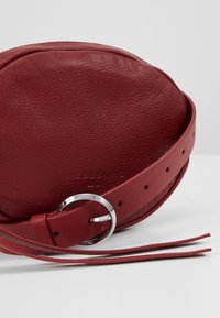 Borsa rotonda in pelle bordeaux con texture, dotata di cinturino in metallo argentato e dettagli frangiati. Presenta il marchio 'LIEBESKIND' impresso.