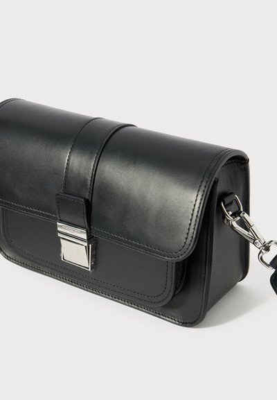 Melna ādas soma ar vāciņu, sudraba rotājumiem un noņemamu ķēdes siksnu. Raksturo gluda tekstūra un šūto detaļu izstrāde.