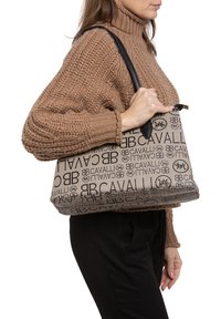 Bruine tote bag met zwarte logo-print, gestructureerde vorm en leren handvatten, gedragen op de arm tegen een achtergrond van een gebreide trui.