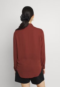 Blouse à manches longues de couleur rouille avec une texture lisse, dotée d'un col et d'un ourlet arrondi. Portée avec un short noir et un accessoire pour queue de cheval.