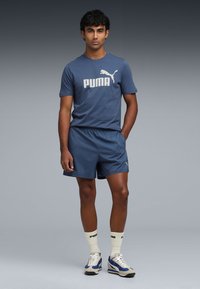 Merenkblue urheiluasu, joka koostuu lyhythihaisesta T-paidasta, jossa on valkoinen Puma-logo, ja yhteensopivista shortseista, yhdistettynä valkoisiin sukkiin ja lenkkareihin.