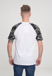 Valkoinen t-paita, jossa on lyhyet camouflage-kuosiset hihat, pyöreä kaula-aukko ja näkyvä kangasrakennetta. Lisäksi on päällä farkut ja kello.