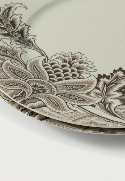 Assiettes en céramique empilées avec des motifs floraux et de feuilles complexes beige et marron sur fond crème, partiellement visibles sur une surface blanche.