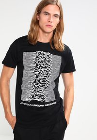 Sort bomulds t-shirt med et hvidt horisontalt linjemønster, der ligner lydbølger, med teksten "JOY DIVISION - UNKNOWN PLEASURES" nederst.