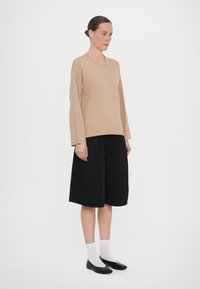 Maglione beige lavorato a maglia con scollatura a V e motivo geometrico, abbinato a pantaloni corti neri fino al ginocchio e scarpe nere basse con calzini bianchi.