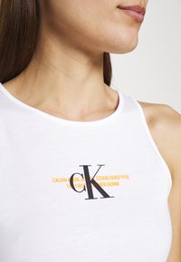 Bílý bezrukávový top vyrobený z bavlny, s velkým černým logem "CK" a oranžovým textem pod ním. Hladká textura a zaoblený výstřih.