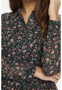 Kwiatowa bluzka z czarnego przezroczystego materiału w wielokolorowe wzory kwiatowe. Posiada klasyczny kołnierzyk i guziki na rękawach.