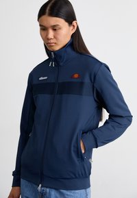 Modra zip-up jakna iz gladkega blaga z visokim ovratnikom, z značilnim črtastim dizajnom in logotipom na prsih.
