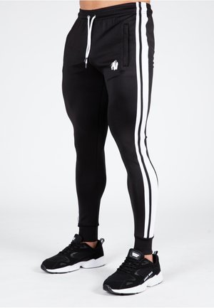 Jogging noir avec des rayures blanches sur les côtés, taille élastique et un petit logo sur la cuisse gauche. Fabriqué dans un matériau lisse et extensible.