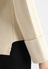 Gestreifter cremefarbener Strickpullover mit einem umgeschlagenen Bündchen und seitlichem Schlitz, strukturierter Stoff sichtbar, kombiniert mit schwarzem Stoff darunter.