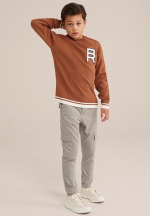 Bruine gebreide trui met geribde manchetten en zoom, voorzien van een witte "R" patch; lichtgrijze cargobroek met meerdere zakken; witte sneakers.