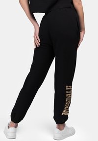 Black sweatpants with a relaxed fit, featuring side pockets and elastic cuffs, adorned with a gold "CONSULT" print on the left leg.