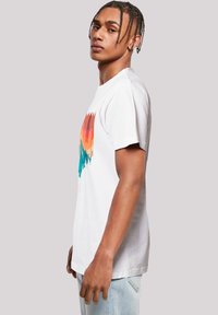 White cotton t-shirt with short sleeves featuring a multicolored graphic of trees and mountains in shades of orange, teal, and brown.