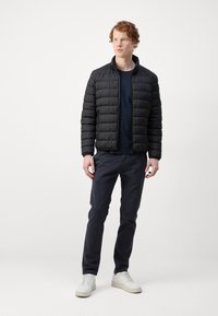 Black puffer jacket with a high collar, textured quilted design. Worn over a navy shirt, paired with dark pants and white sneakers.