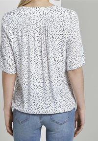 Weiße Bluse mit schwarzem und blauem Punktmuster, kurze Ärmel, gerafftes Rückendetail, weicher Stoff, kombiniert mit hellblauen Jeans.