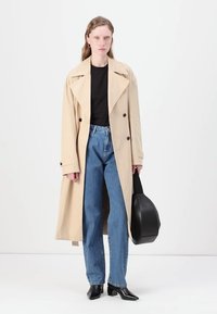 Beige dobbelbrettet trenchcoat med brede krager, brukt over en svart topp og blå høytlivsjeans. Svart spiss tå-sko og håndveske.