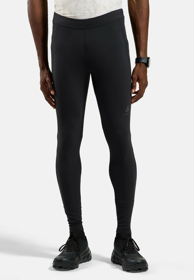 Homme portant un legging de sport noir ajusté, des chaussures de course noires, un haut blanc et une montre de sport noire, debout sur un fond blanc.