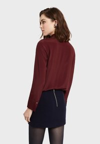 Bordowa bluzka z długim rękawem o luźnym kroju, zestawiona z granatową spódnicą z tylnym zamkiem. Widoczny jest teksturowany materiał oraz mankiety zapinane na guziki.