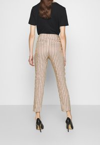 Beżowe spodnie w paski z pionowymi czerwonymi liniami, wysokim stanem, zwężanymi nogawkami i teksturowanym materiałem. Połączone z czarnymi butami na obcasie.
