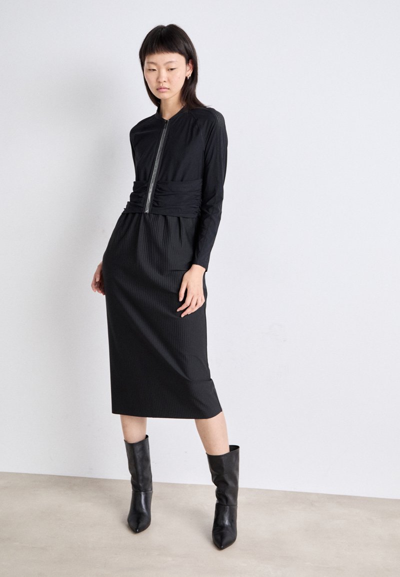 Schwarzes Kleid mit langen Ärmeln, tailliertem Oberteil und geraffter Taille, das in einen Midilängenrock mit vertikalen Rippungen übergeht. Getragen mit schwarzen Ankle Boots.