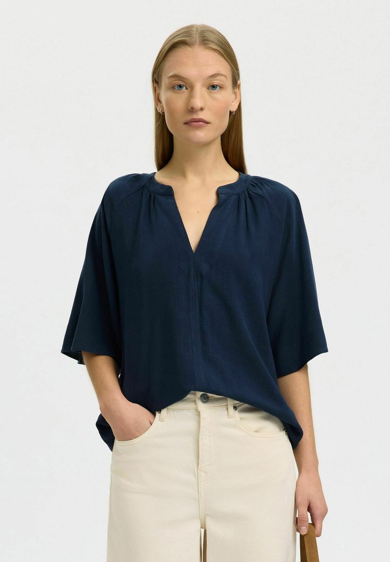 Femme aux cheveux blonds raides portant un chemisier bleu marine ample à col en V et un pantalon crème taille haute, une main dans la poche et une expression neutre.