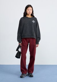 Felpa nera oversize con una grafica a forma di cuore, abbinata a pantaloni a gamba dritta bordeaux. Il modello tiene una piccola borsa nera.