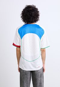Camiseta deportiva blanca con acentos en azul, verde y rojo. Cuenta con una parte trasera de malla texturizada y un patrón a cuadros. Corte suelto con mangas cortas.