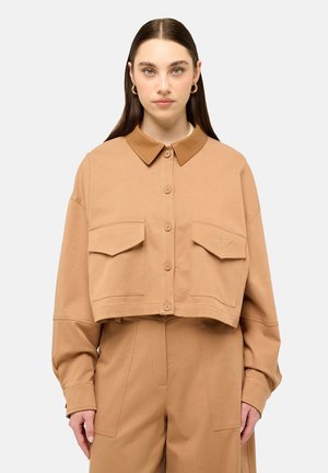 Donna con capelli lunghi e scuri che indossa una giacca corta color sabbia con grandi tasche frontali e pantaloni coordinati a vita alta, in piedi davanti a uno sfondo chiaro.