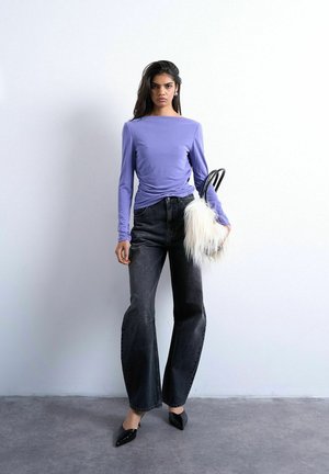 Donna che indossa un top a maniche lunghe viola, jeans neri a gamba larga, scarpe nere con punta, tiene in mano una borsa bianca e pelosa, in piedi contro un muro bianco.