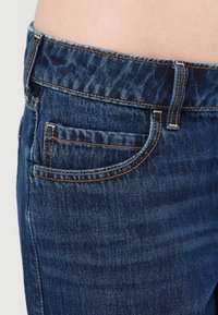 Μπλε τζιν παντελόνι με εφαρμοστή σχεδίαση, εμφανείς ραφές σε πορτοκαλί και λεπτομέρεια μπροστινής τσέπης. Το υλικό φαίνεται υφασμάτινο.