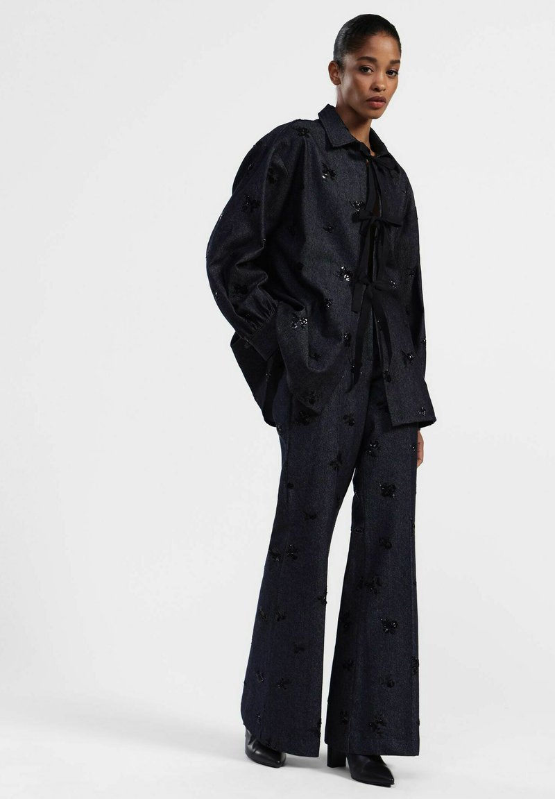 Completo in denim nero composto da un giubbotto ampio con decorazioni floreali e pantaloni a campana abbinati. Include un nastro da annodare al colletto.