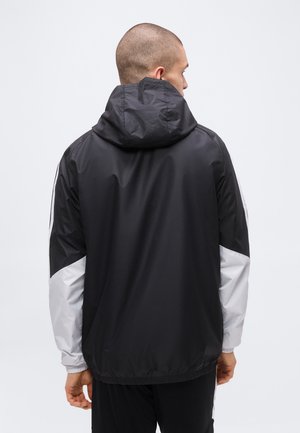 Mann trägt schwarze und graue Kapuzen-Windjacke, steht mit dem Rücken zur Kamera vor einem schlichten weißen Hintergrund.
