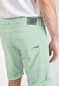 Shorts en denim vert clair avec des revers, comportant deux poches arrière et une patch en cuir noir sur la ceinture.