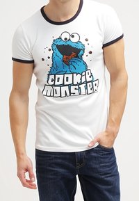 Mężczyzna w białej koszulce z kolorową grafiką Cookie Monstera, trzymający ciastko, w ciemnych dżinsach i w luźnej pozie.