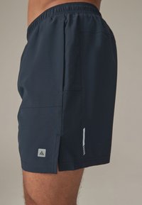 Costume da bagno corto blu navy con fascia elastica in vita, tasche laterali, texture traforata sottile e una piccola patch con logo sul lato sinistro.