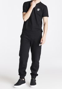 Zwarte katoenen t-shirt met een klein wit logo op de borst, gecombineerd met bijpassende zwarte joggers. Inclusief zwarte sneakers met witte zolen.