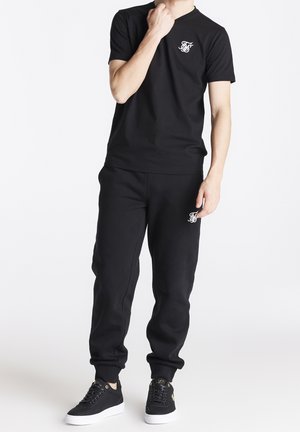 T-shirt en coton noir avec un petit logo blanc sur la poitrine, associé à un pantalon de survêtement noir assorti. Comprend des baskets noires avec des semelles blanches.