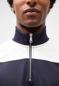 Polo mezza zip blu navy e bianco con colletto alto, dotato di una cerniera argentata e tessuto testurizzato. Vista ravvicinata del colletto.