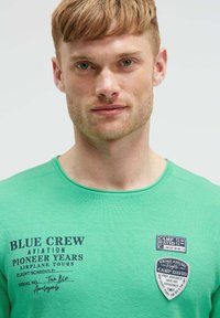 Grünes, strukturiertes Langarmhemd mit bedrucktem Text und grafischen Patches. Rundhalsausschnitt, figurbetonte Passform, lässiges Design.